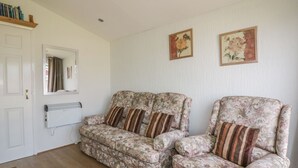 A living room with two floral-patterned sofas, a white radiator, a wooden shelf with books, and two framed floral paintings on the wall. - Warren Cove (Wadebridge)