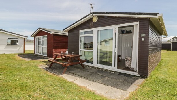 A small, single-story mobile home with a picnic table in front, a sliding glass door, and a number '157' on the door. - Warren Cove (Wadebridge)