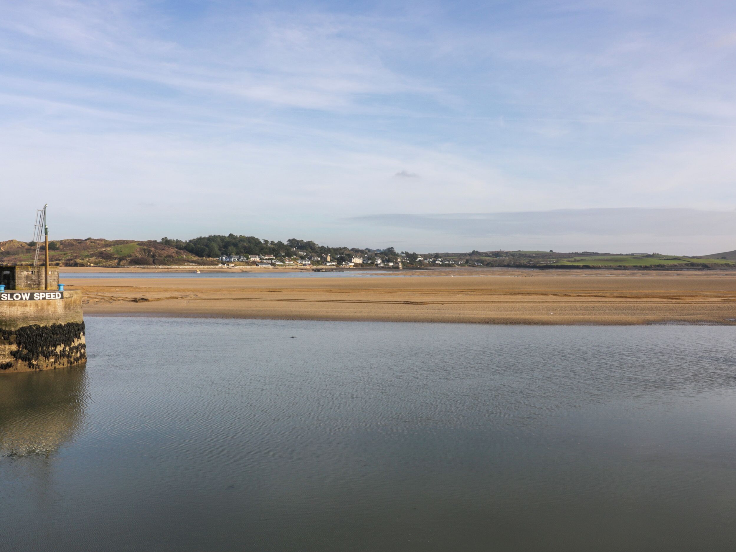 Un paysage côtier avec une plage de sable, une étendue d’eau calme et une ville lointaine, avec des maisons et de la végétation.