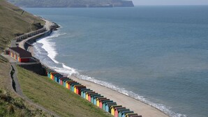 A coastal road with colorful beach huts and a view of the sea and cliffs. - Flying Scotsman @ Engine Shed (Whitby)