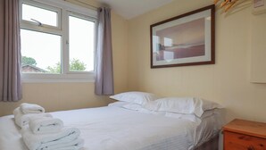 A neatly made bed with white linens and towels, a window with curtains, a framed picture on the wall, and a wooden nightstand. - Pepper Cove (Wadebridge)