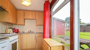 A kitchen with wooden cabinets, a red curtain, a window, and a view of a backyard. - Pepper Cove (Wadebridge)