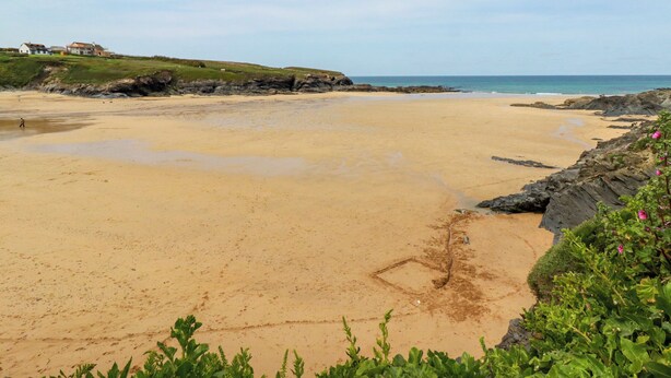 A beach with a rocky shoreline, a grassy hill with a building, and clear skies. - Pepper Cove (Wadebridge)