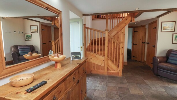 Un escalier en bois menant à un deuxième étage, un sol carrelé en pierre, un comptoir en bois avec un bol et une télécommande, et un fauteuil en cuir.