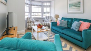Un salon avec un canapé de couleur turquoise, des fauteuils à rayures, une table basse sur laquelle se trouve un panier de fruits et un meuble pour la télévision.