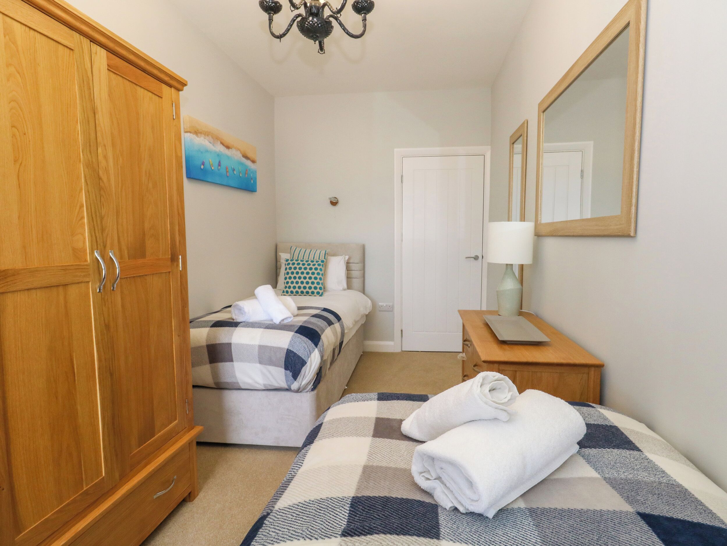 A bedroom with a large wooden wardrobe, a bed with a checkered bedspread, a bedside table with a lamp, and a painting on the wall.