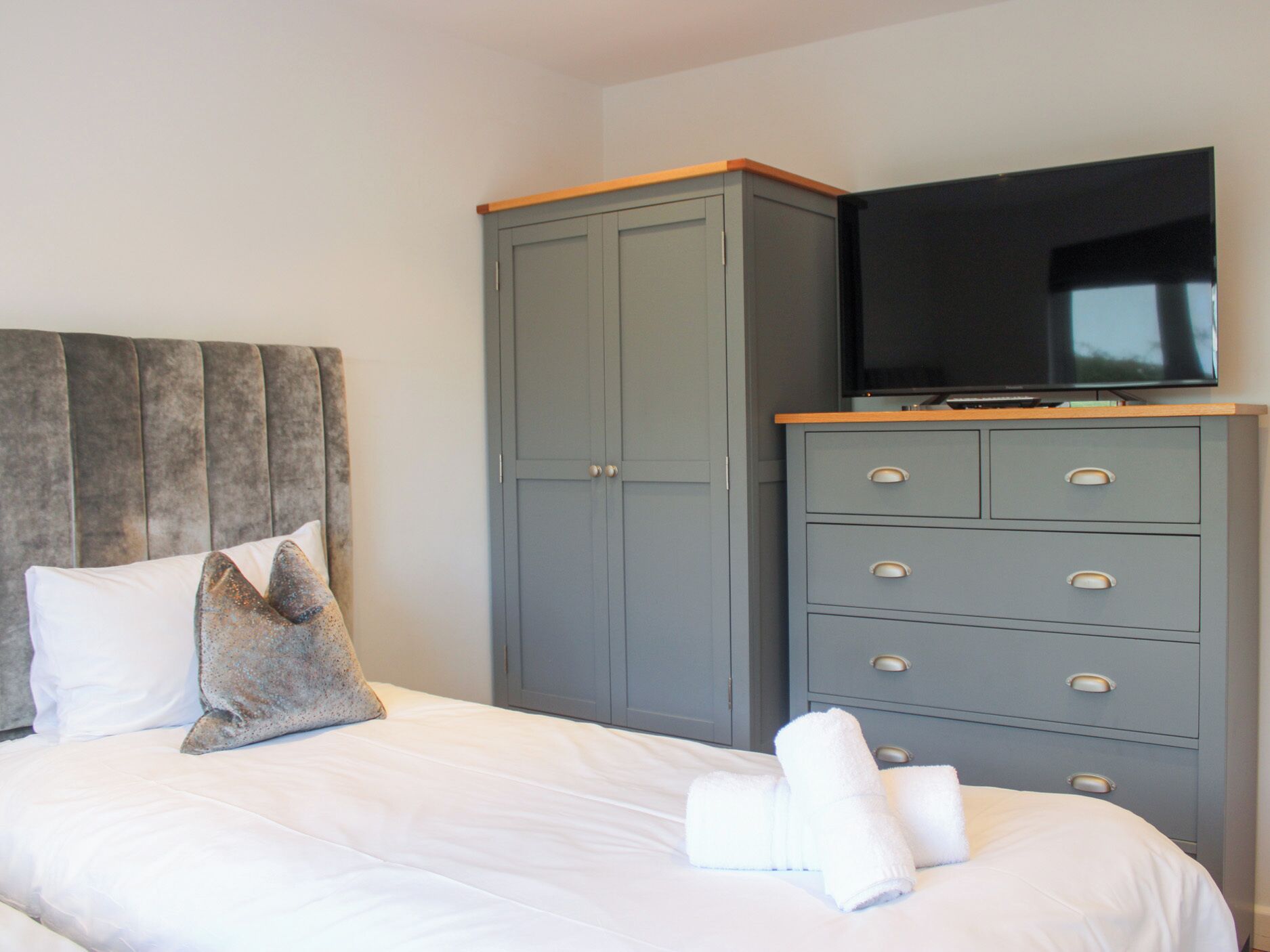 A hotel room with a bed, a grey wardrobe with drawers, a television on top, and a pillow on the bed.