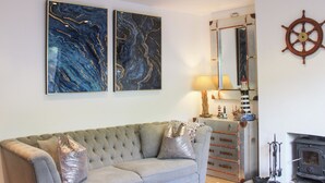 A living room with a grey sofa, two abstract paintings, a wooden ship's wheel, a mirror, and a fireplace. - Old Police House (Salcombe)