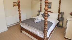 A four-poster bed with a herringbone-patterned bedspread, a white towel on the bed, and a nightstand with a lamp and a small table with a vase of flowers. - Clearbrook Lodge (Carmarthen)