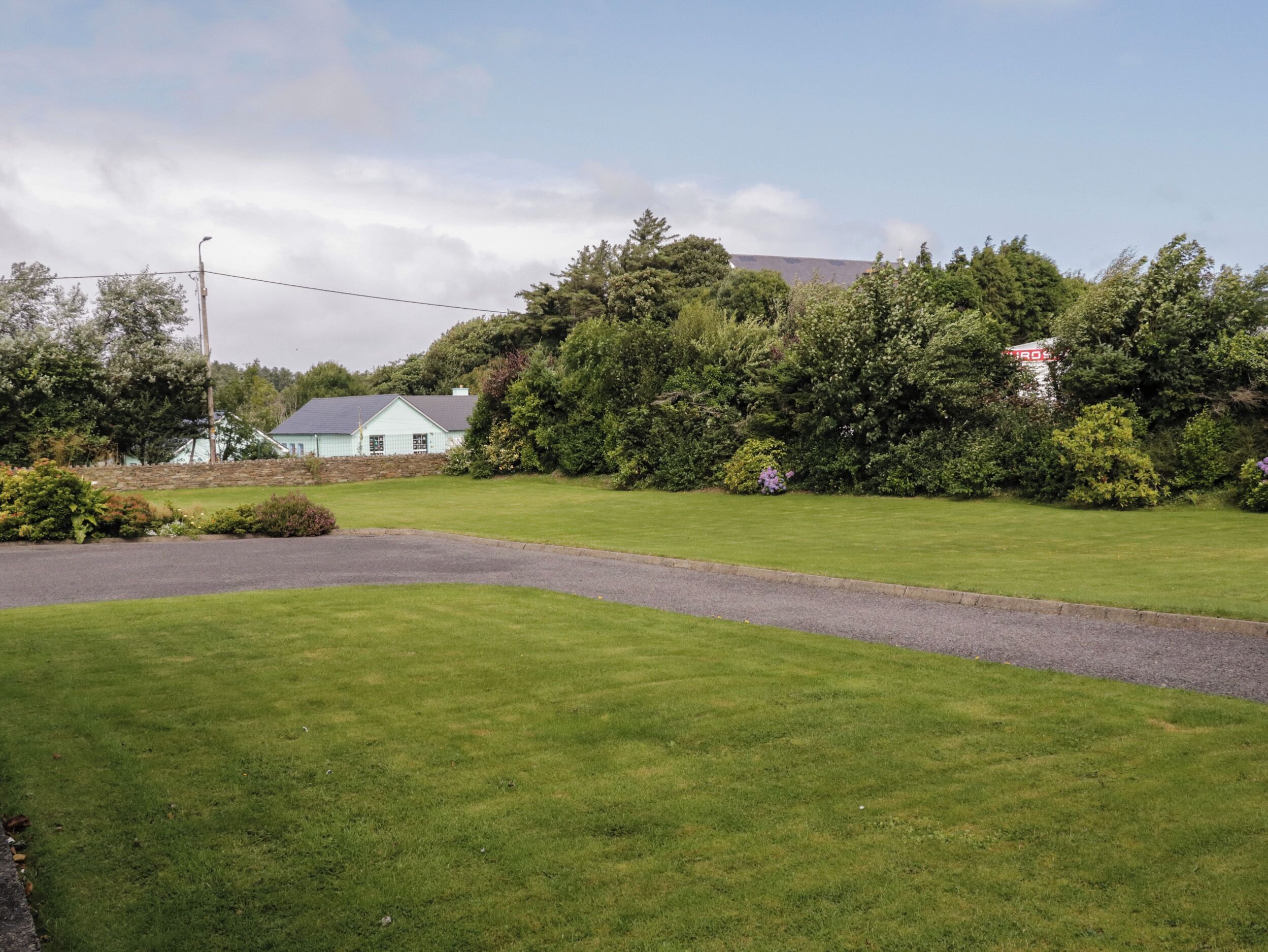 A grassy area with a paved path, a stone wall, and a building with a light blue roof.