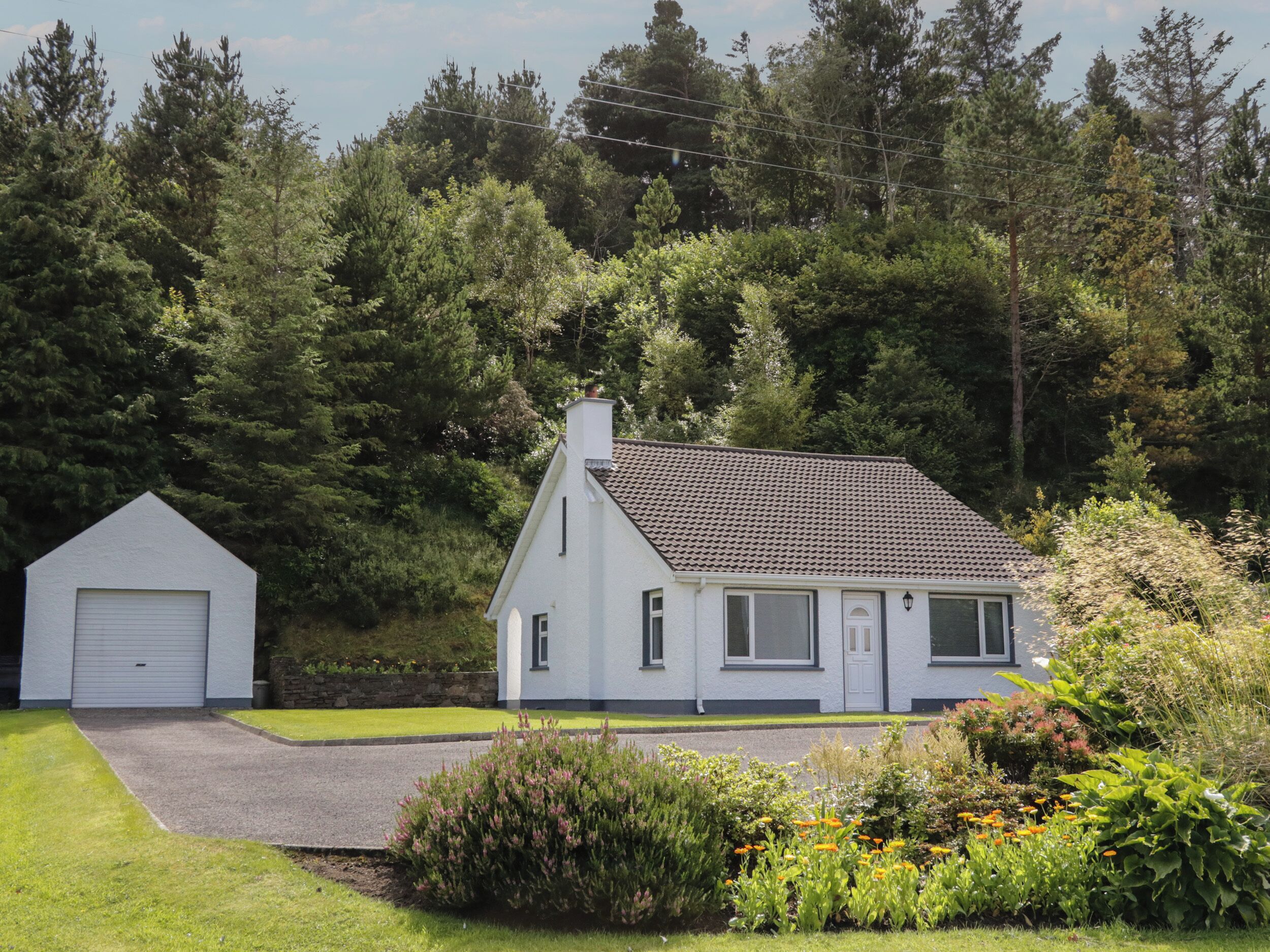 A white house with a tiled roof, a garage, and a well-maintained garden with various plants and flowers.
