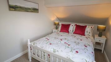 A bedroom with a white bed, floral bedding, two red pillows, two bedside lamps, and a landscape painting on the wall.