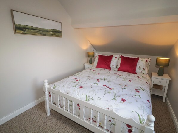 Une chambre à coucher avec un lit blanc, une literie à motifs floraux, deux oreillers rouges, deux lampes de chevet et un tableau représentant un paysage accroché au mur.