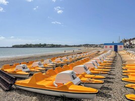 Una hilera de botes amarillos alineados en una playa pedregosa, con vista despejada al mar y una costa lejana con edificios.