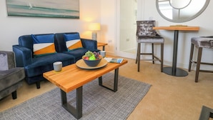 A modern living room with a blue sofa, a wooden coffee table with fruit, a round mirror, and a glass door leading to a balcony.