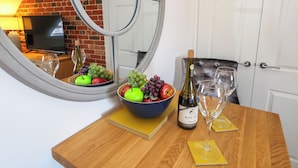 A wooden table with a bowl of fruit, a bottle of wine, and two wine glasses. A mirror reflects a brick wall and a television in the background.