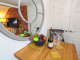 Una mesa de madera con un cuenco de fruta, una botella de vino y dos copas de vino. Un espejo refleja un muro de ladrillo y un televisor al fondo.