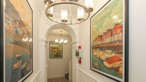 A hallway with framed artwork depicting coastal scenes, a chandelier, and a patterned floor.
