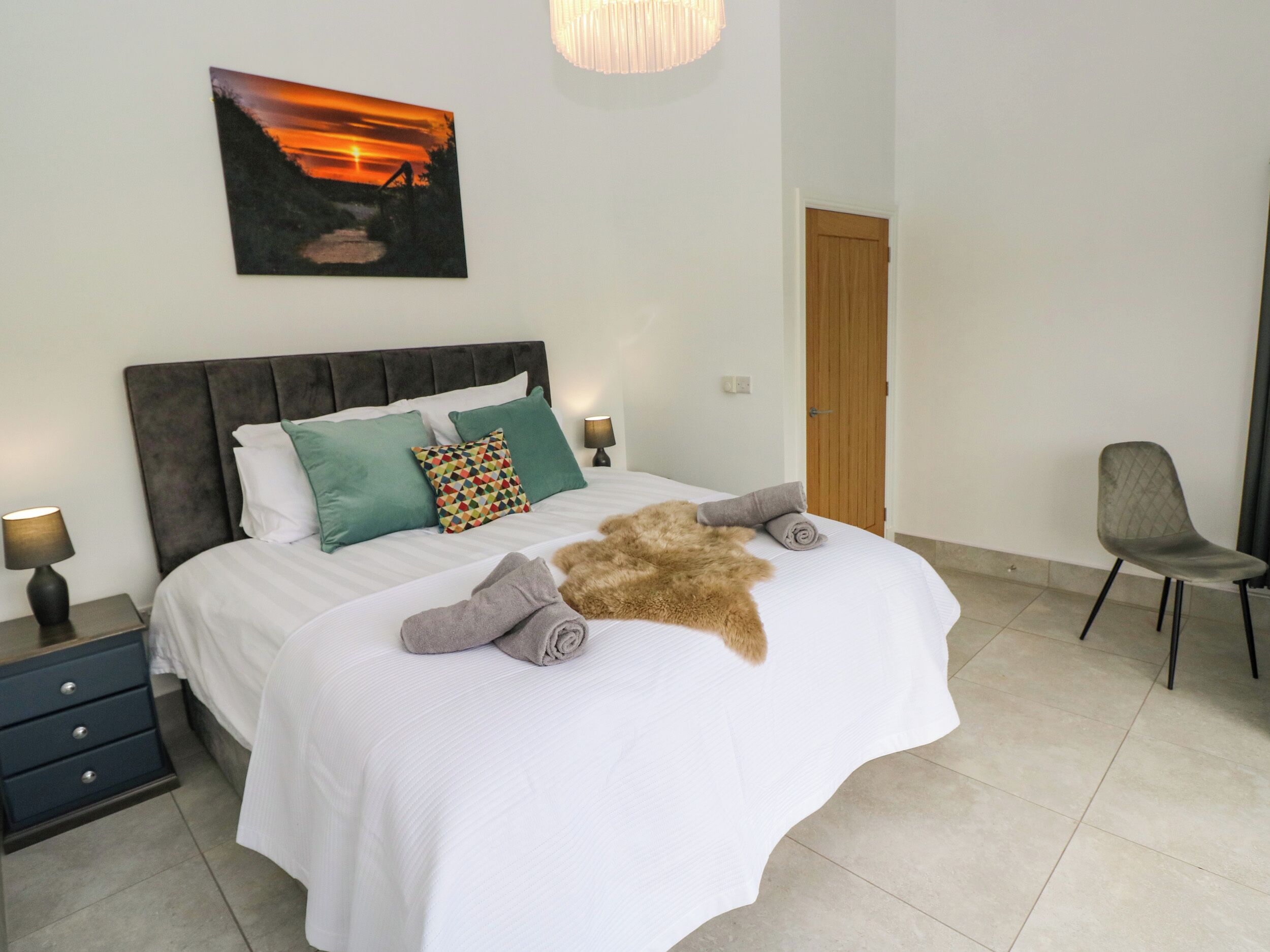 A neatly made bed with white linens and multiple pillows, a wooden headboard, a wall-mounted painting of a sunset, a small table with a lamp, and a chair.
