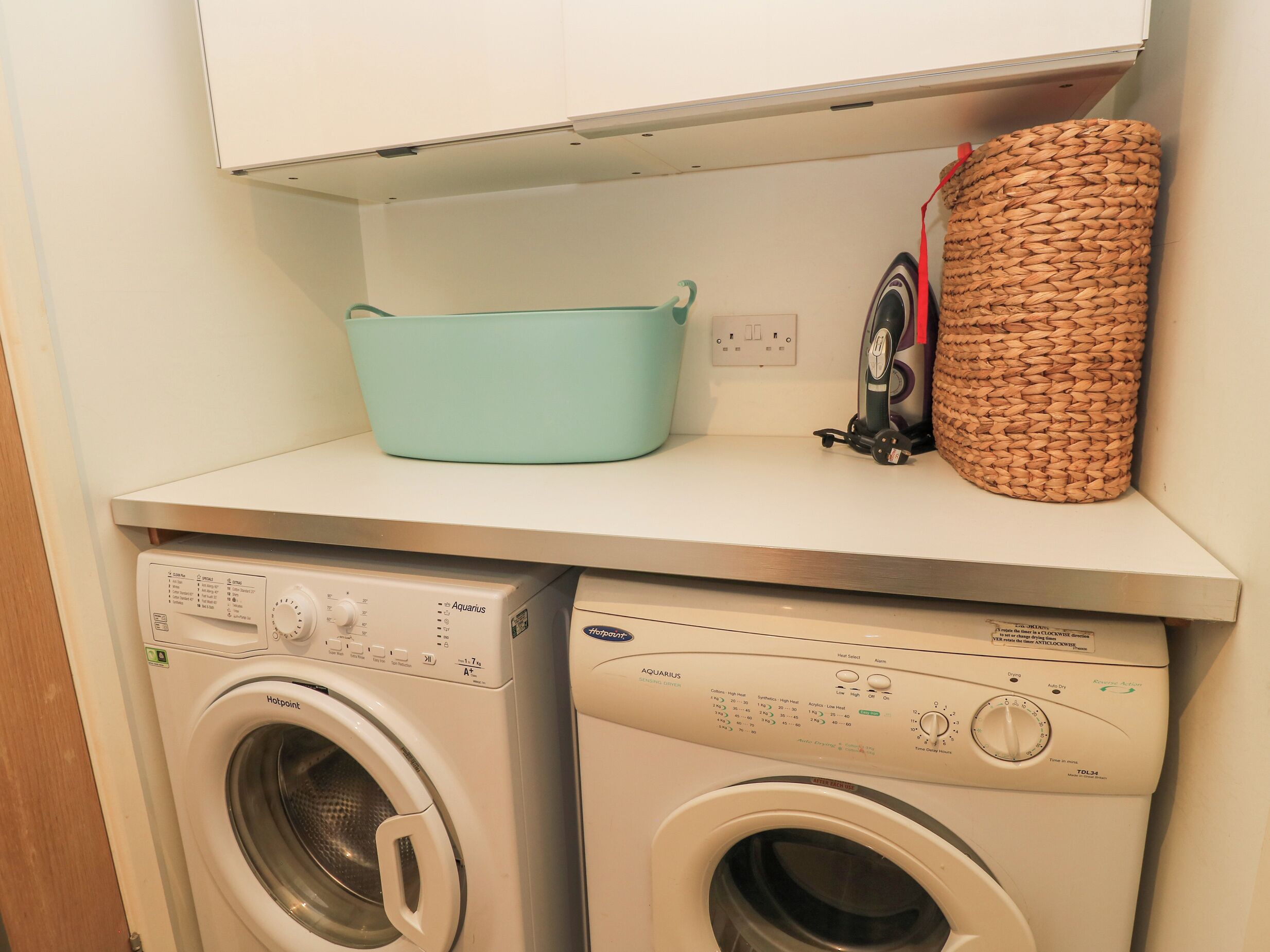 Un espace buanderie comprenant deux machines à laver, un évier de couleur turquoise et un panier en osier.