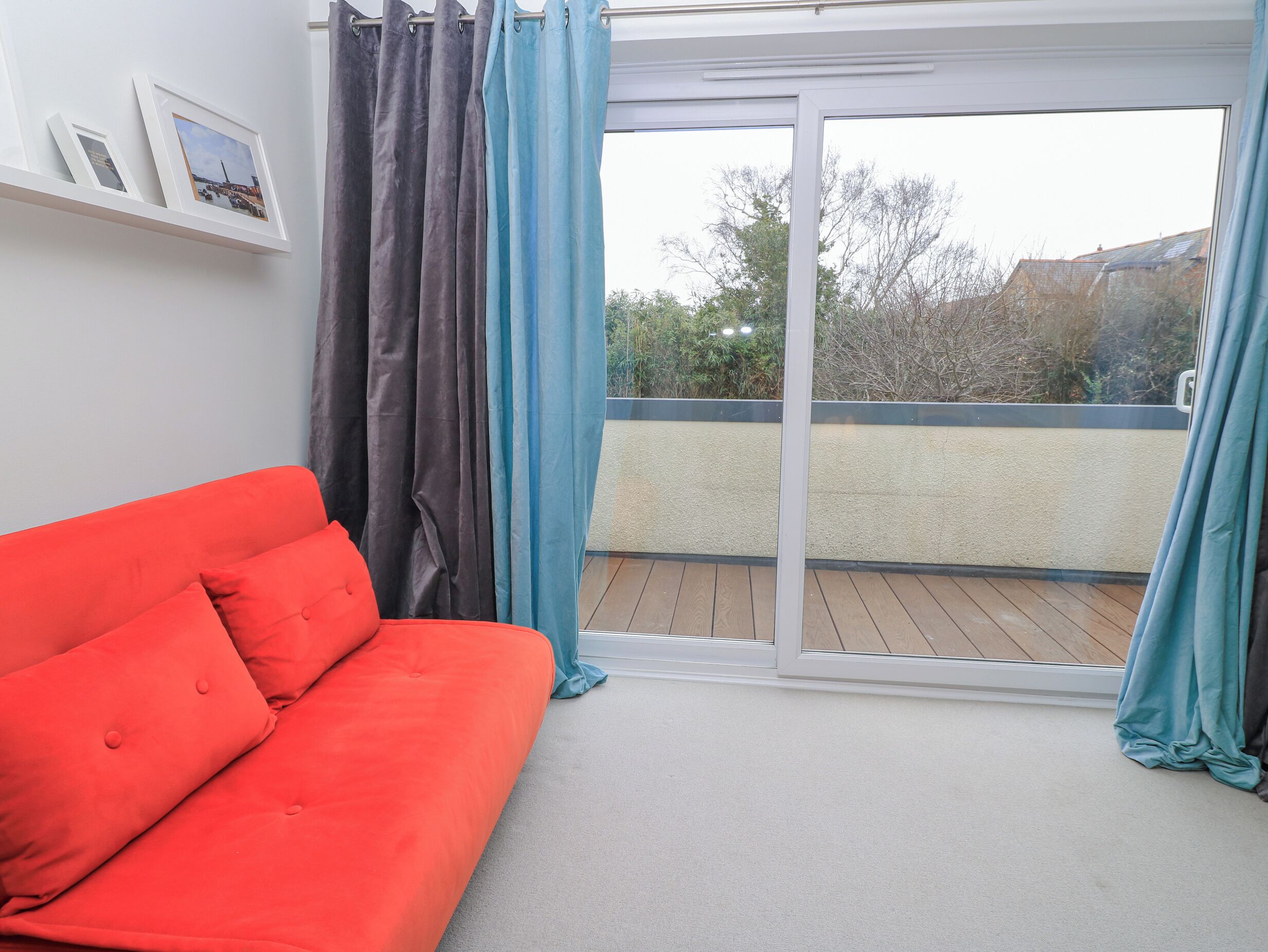 A room with a red sofa, a sliding glass door to a balcony, and a wall-mounted shelf with framed pictures.