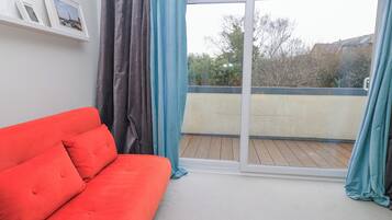 A room with a red sofa, a sliding glass door to a balcony, and a wall-mounted shelf with framed pictures.