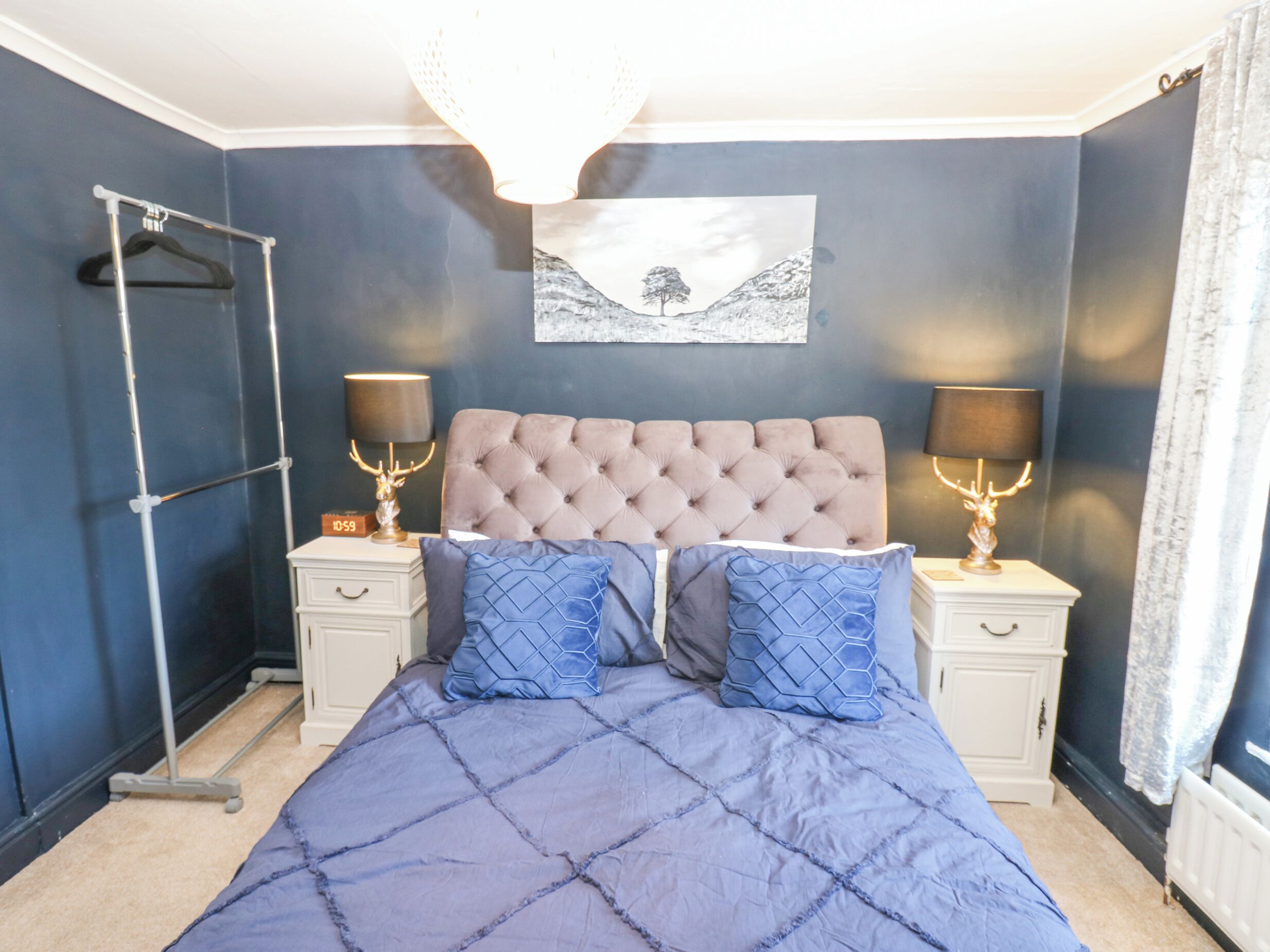 Un dormitorio con cabecera capitonada, dos mesitas de noche con lámparas, cama con colcha azul y una obra de arte colgada en la pared.