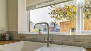 A kitchen with a large white sink, a window with a view of a wooden fence and trees, and two potted plants on the windowsill. - Farmer's Rest (Ely)