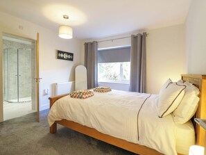 A bedroom with a bed, a bedside table, a window with curtains, a radiator, and a door leading to a shower area. - Farmer's Rest (Ely)