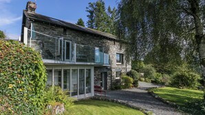 Une maison moderne en pierre, dotée d’une véranda en verre, entourée d’une végétation luxuriante et accessible par une allée en gravier.