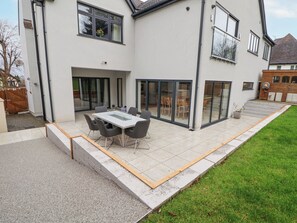 Une maison moderne dotée d’un patio comprenant une table et des chaises, de grandes portes-fenêtres coulissantes et une pelouse bien entretenue.