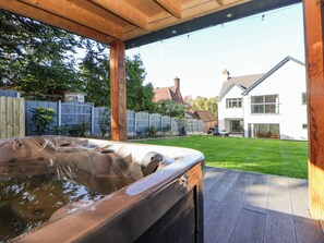A hot tub in a covered outdoor area with a wooden deck, a lawn, and a white house in the background. - Hafan Y Mor (Prestatyn)