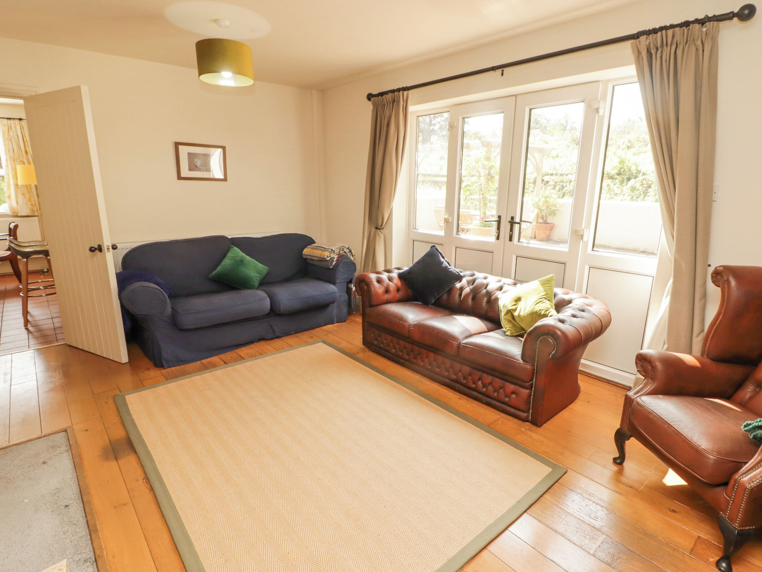 A living room with a blue sofa, a brown leather sofa, and a brown leather armchair. There is a large window with curtains, a door leading to another room, and a framed picture on the wall.