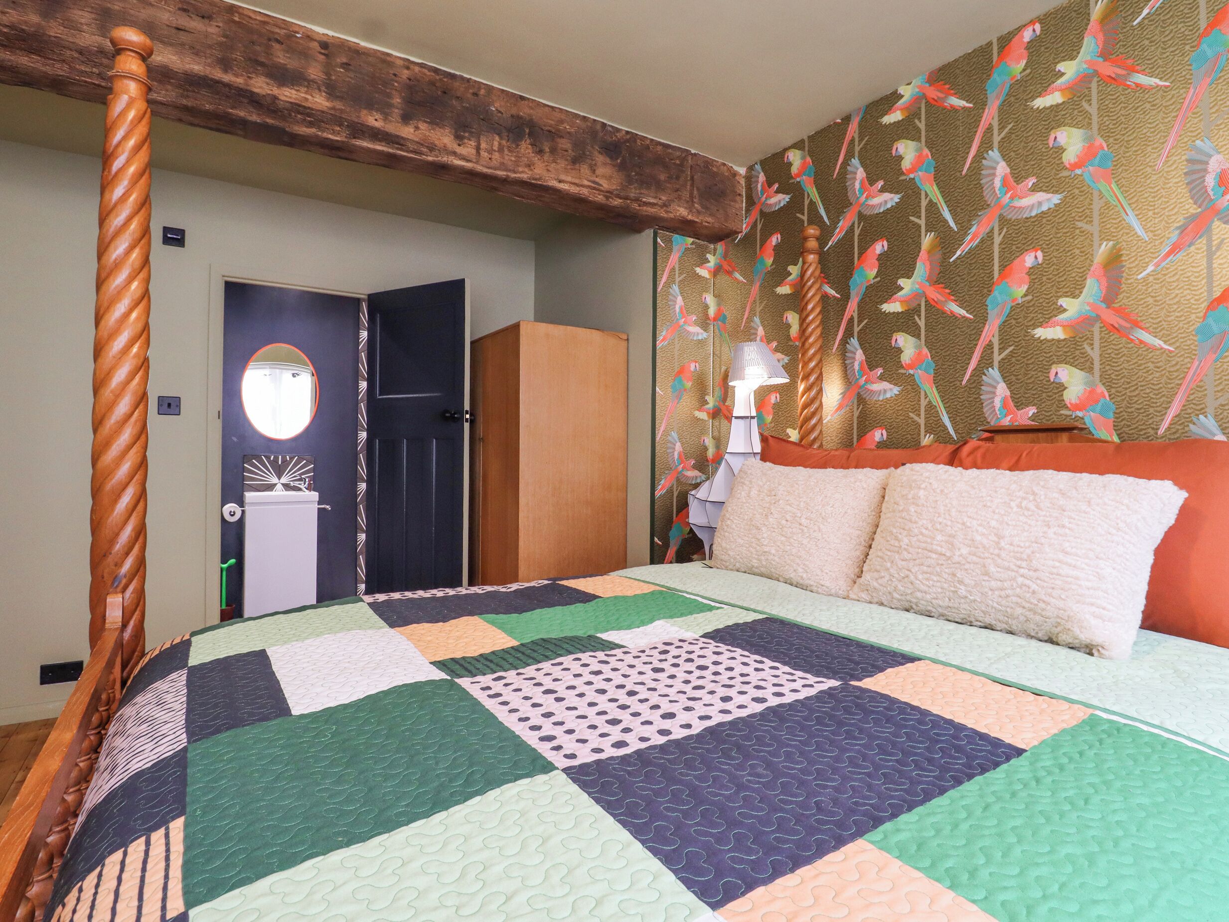 Une chambre à coucher avec un lit à baldaquin en bois, une courtepointe aux couleurs vives, une armoire et un mur orné d’un motif de perroquets.