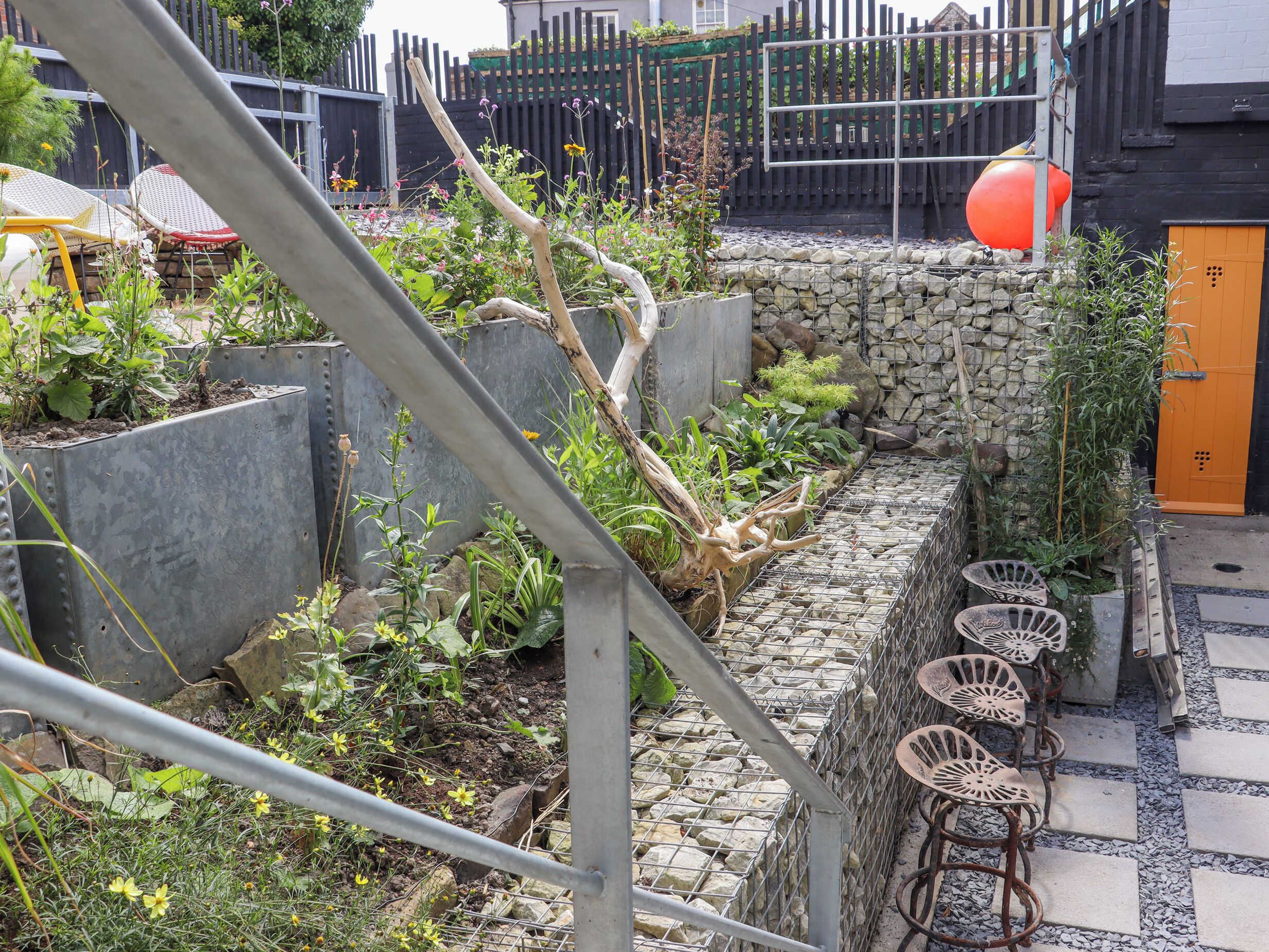 A garden with a stone wall, metal chairs, and a wooden fence.