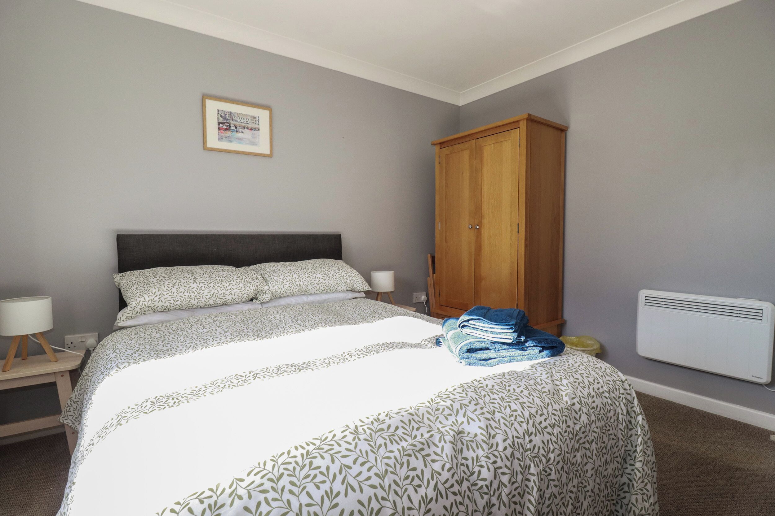 Une chambre avec un grand lit, une armoire en bois, une table de chevet avec une lampe et un tableau encadré au mur.