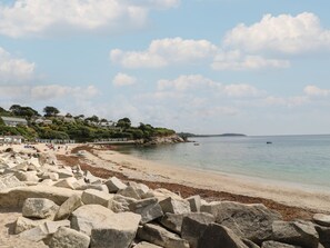 A coastal beach with large rocks in the foreground, a sandy shore, and houses on a hillside in the background. - Coombe Cottage (Falmouth)