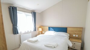 Ein ordentlich bezogenes Bett mit weißen Laken und zwei gefalteten Handtüchern, ein Nachttisch mit Lampe, ein hölzernes Kopfteil und ein Fenster mit blauen Vorhängen.