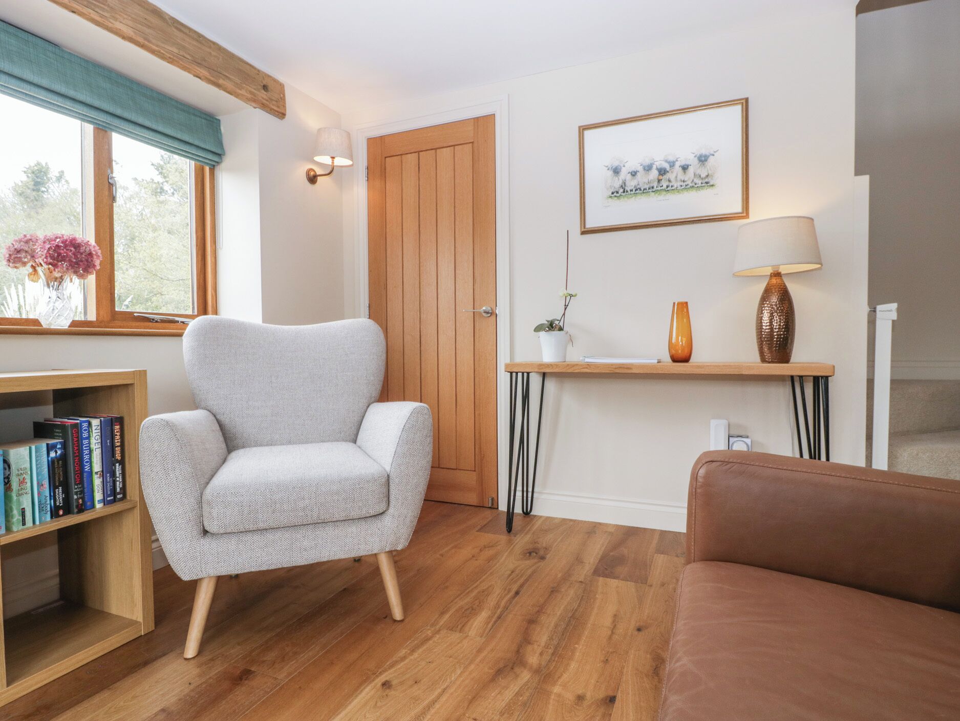 Ein Wohnzimmer mit grauem Sessel, braunem Sofa, einer Holzregalanlage, einer Holztür, einem gerahmten Bild, einem kleinen Tisch mit Lampe und einem Fenster mit Vorhang.