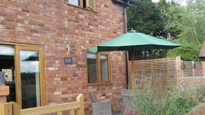 A brick building with a wooden gate, a green umbrella, and a flower bed with red and purple flowers. - The Cartshed (Worcester)
