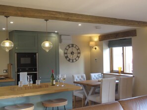 A modern kitchen with a central island, dining area with white upholstered chairs, and a wall clock. - The Cartshed (Worcester)