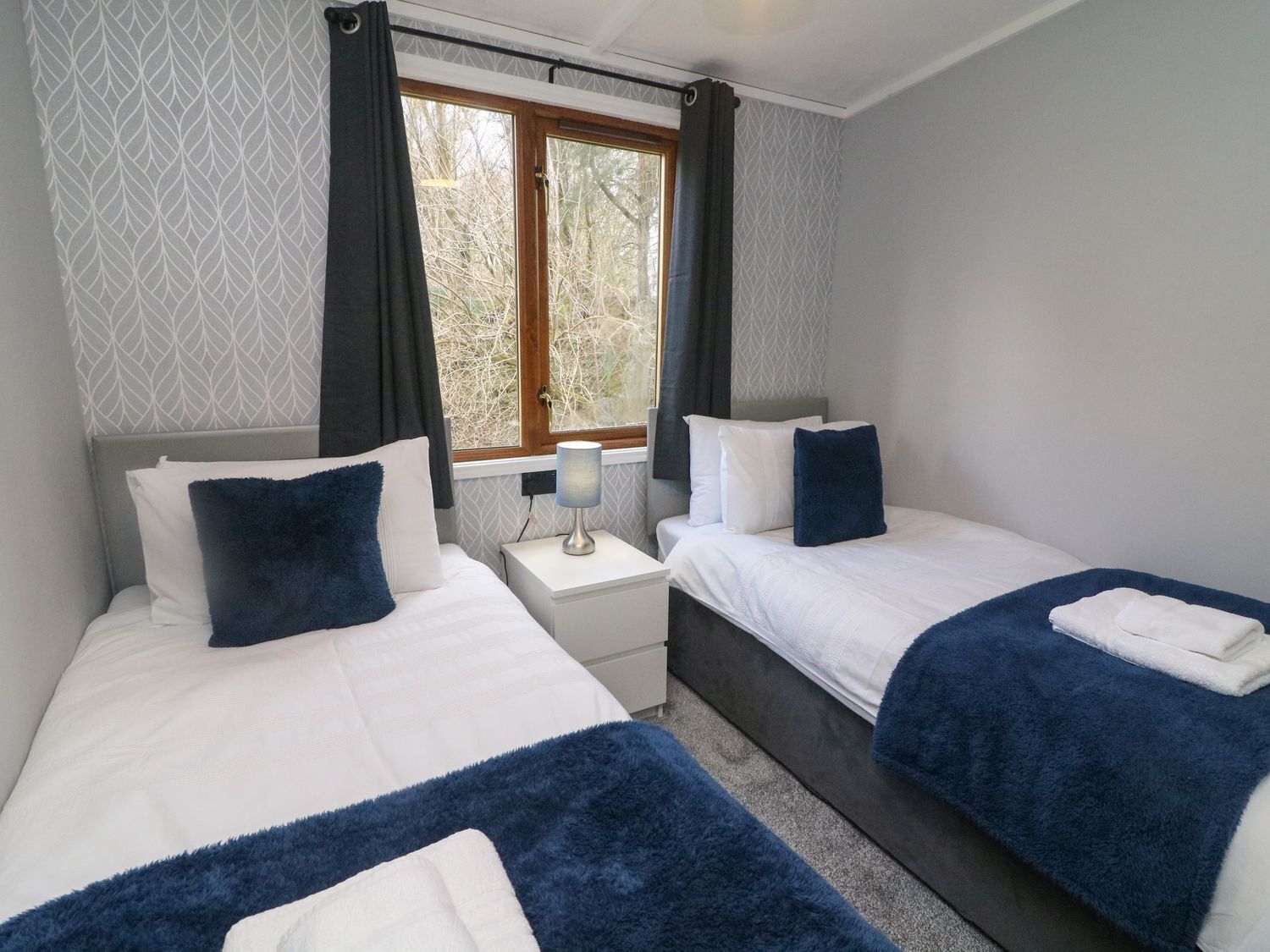 A bedroom with two beds, a window with curtains, a bedside table with a lamp, and patterned wallpaper.