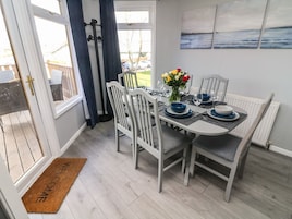 Un comedor con una mesa dispuesta para cuatro, sillas, un florero con flores y vistas al exterior a través de una ventana.
