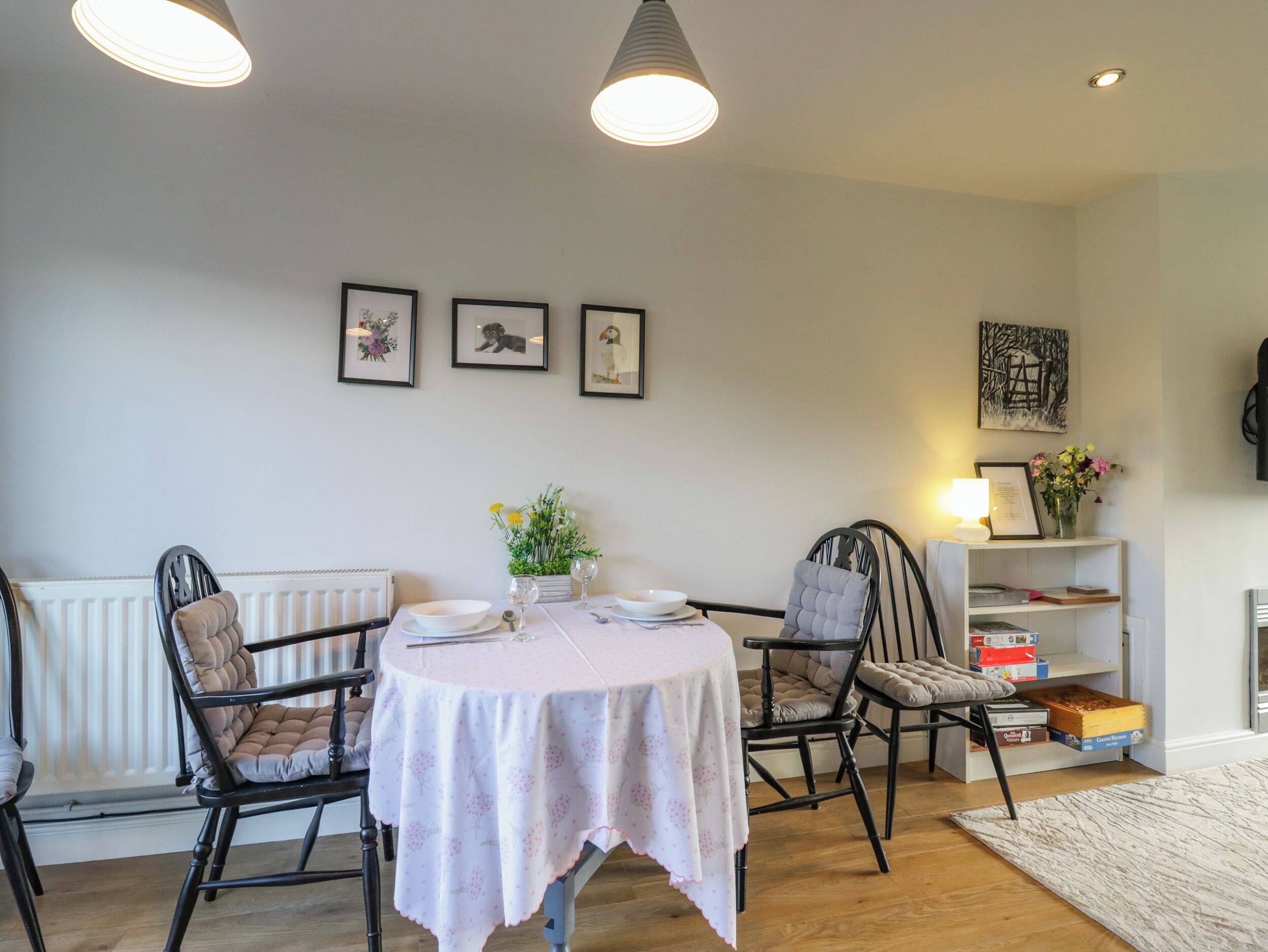 Un comedor con una mesa redonda, cuatro sillas y un mantel blanco. En la pared hay cuadros enmarcados, una estantería con libros y un jarrón de flores, y un radiador debajo de la mesa.