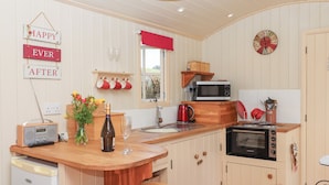 A compact kitchen with a wooden bar counter, a microwave, a kettle, and a small oven. There are three stools at the bar, a wine bottle, and a radio.