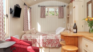 A cozy room with a bed, a desk, a chair, a small table with a bottle and glass, and a window with curtains.