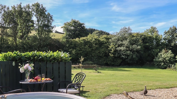 A hot tub surrounded by outdoor furniture, a grassy lawn, and a wooden fence with greenery.