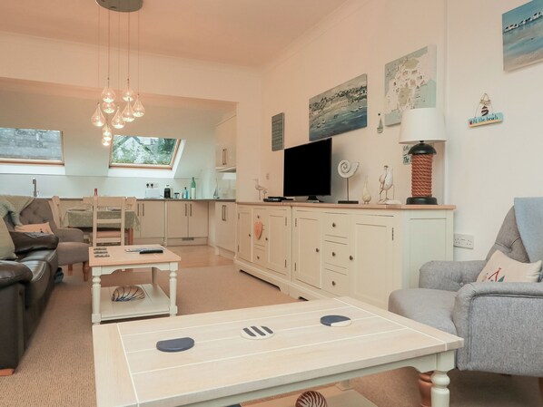 A modern living room with a sofa, a coffee table, and a television. There is a kitchen area with cabinets and a dining table. - 22 Bolt Head (Salcombe)