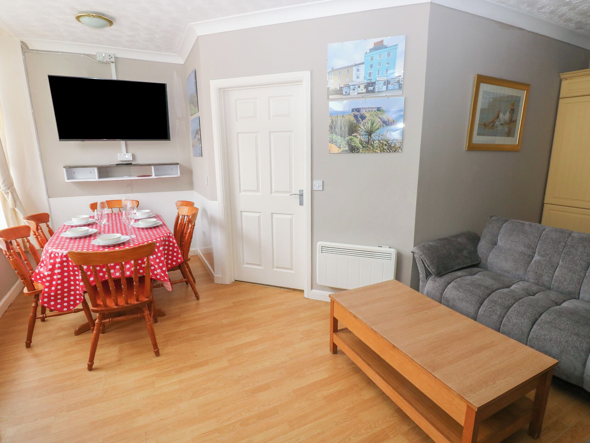 A living room with a dining table set for four, a grey sofa, a wooden coffee table, a flat-screen TV mounted on the wall, and a radiator.