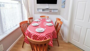 A dining area with a table set for six, wooden chairs, a TV mounted on the wall, and a window with curtains. - The Hilton Retreat (Tenby)